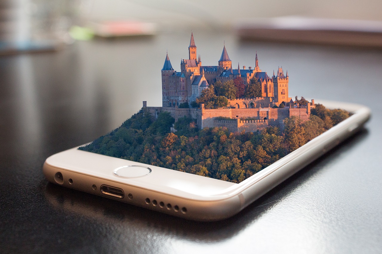 Ein Smartphone liegt auf einem dunklen Tisch. Auf dem Display scheint eine realistische 3D-Landschaft mit einer großen mittelalterlichen Burg auf einem bewaldeten Hügel aus dem Gerät herauszuragen. Die Szene wirkt wie eine digitale Erweiterung und symbolisiert virtuelle Welten oder digitale Erlebnisse der Stadt-App.